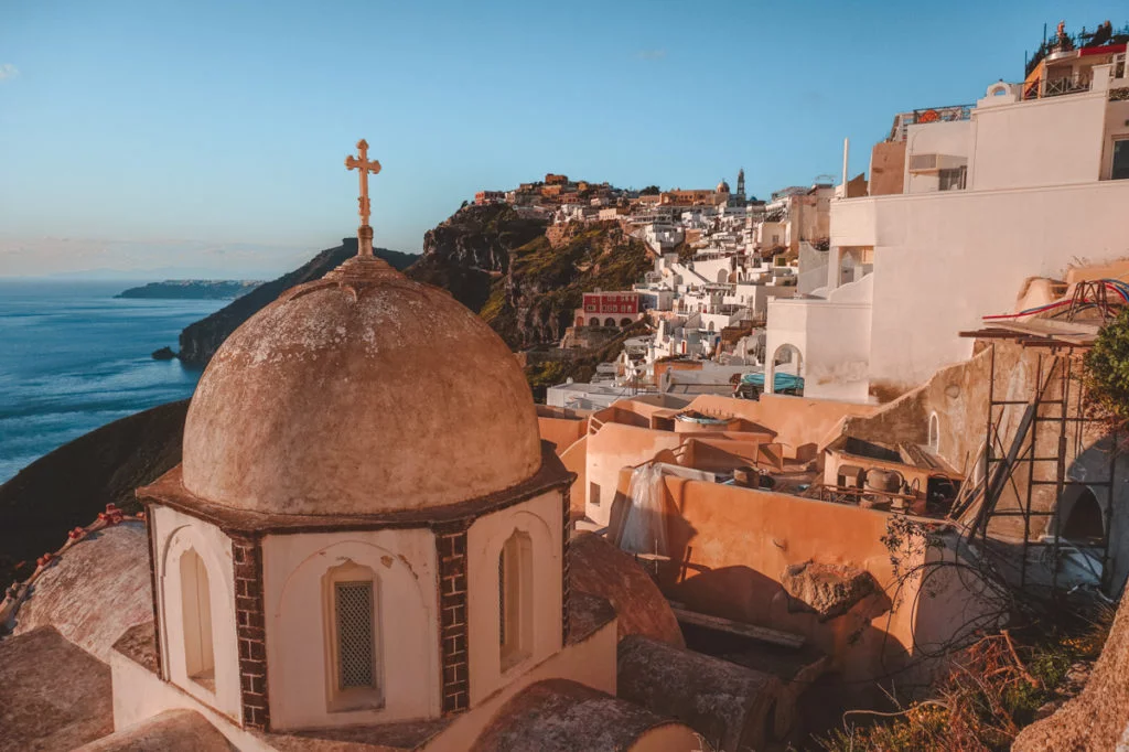 Sunset on the island of Santorini with a church and its rooftop cross standing tall in the foreground, while Fira and Santorini's famous volcanic landscape glows bright in the background. 