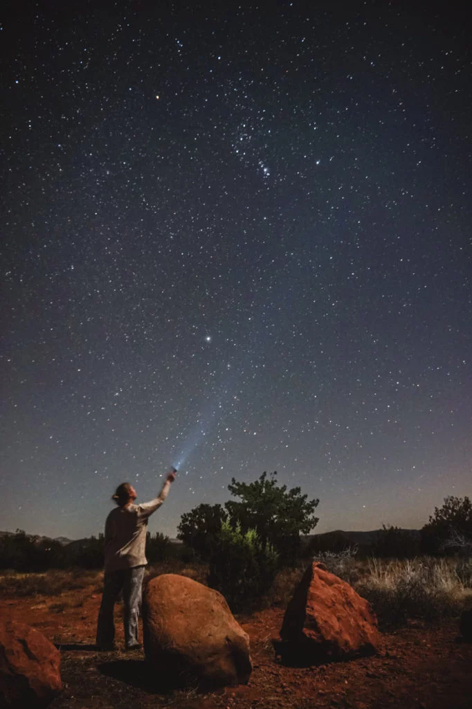 Standing in the middle of a red rock landscape, shining a flashlight up at the glittering stars. 