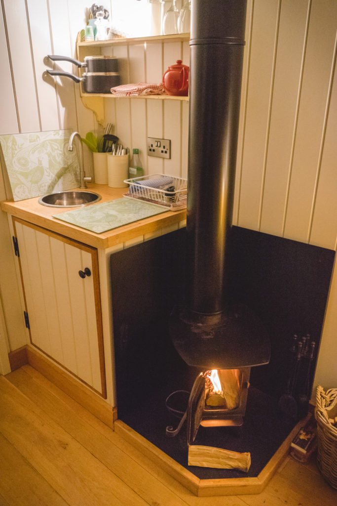 A photo of the fire in the log burning stove and the sink / kitchen space behind it. 